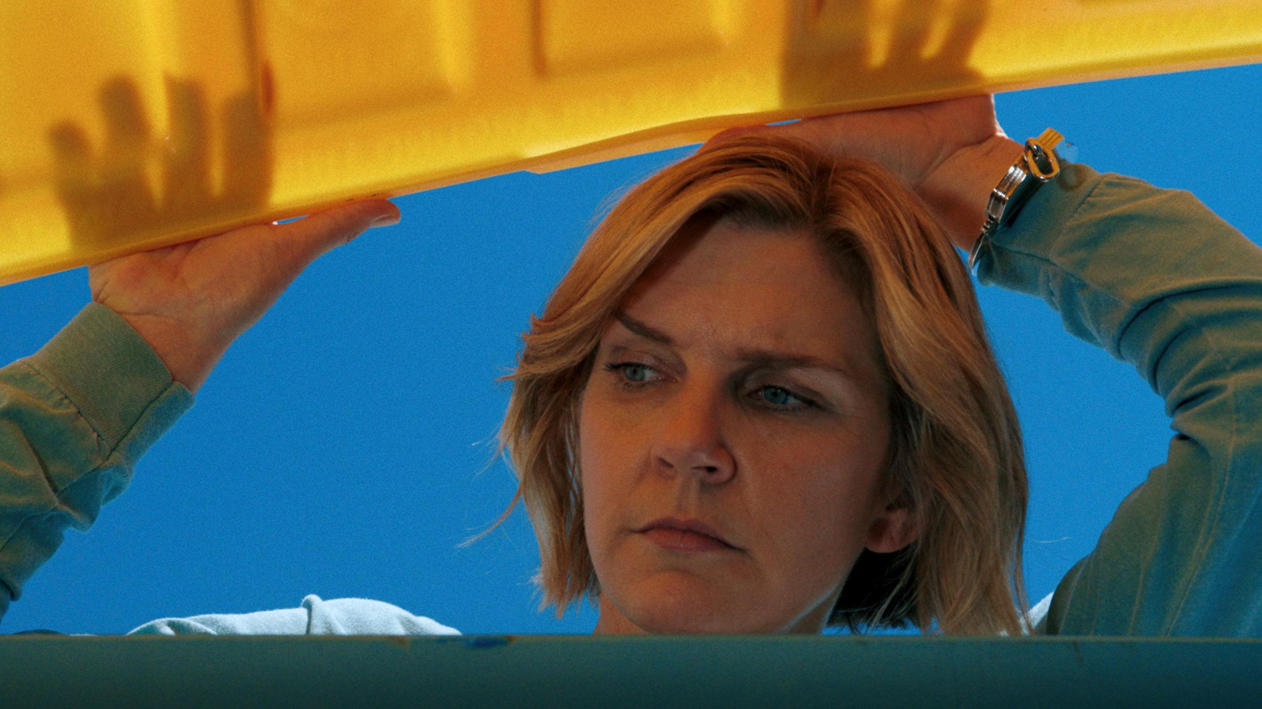 Rhea Seehorn seen from below opening a large yellow lid of a large container and looking inside, in a still from Pluribus