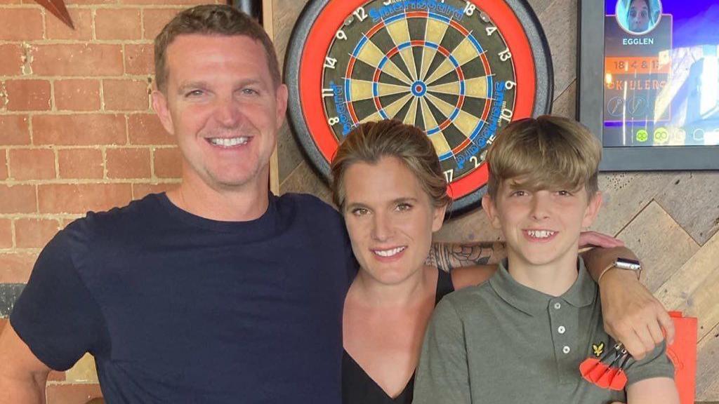 A man, a woman and a teenage boy smiling for a photo. They have their arms around each other and are standing in front of a dart board. The man is holding three darts in his left hand.