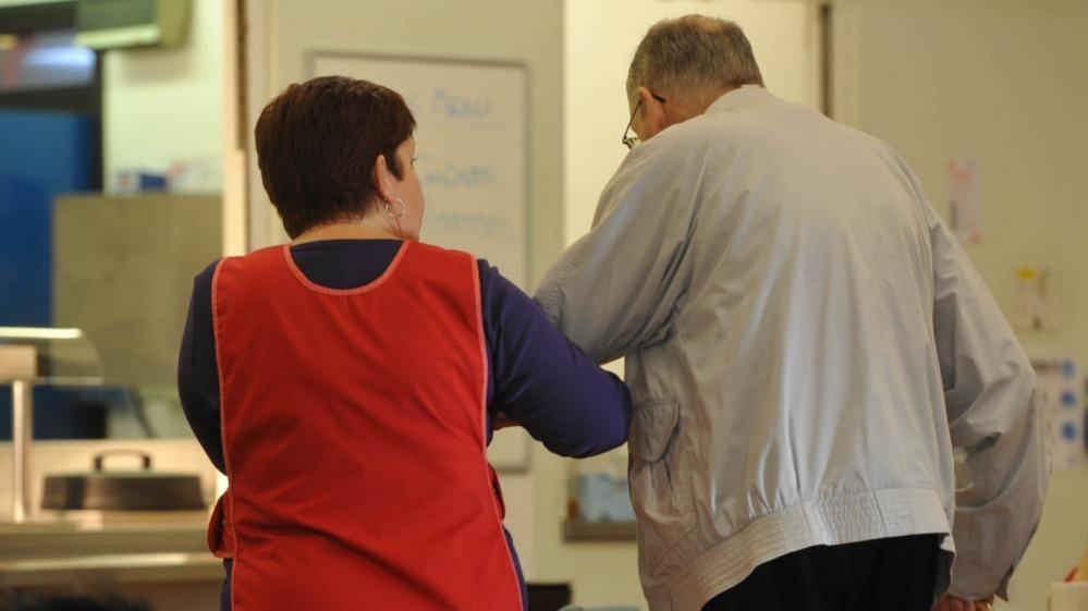 A female care worker wearing a red tabard helps an older man in a grey jacket