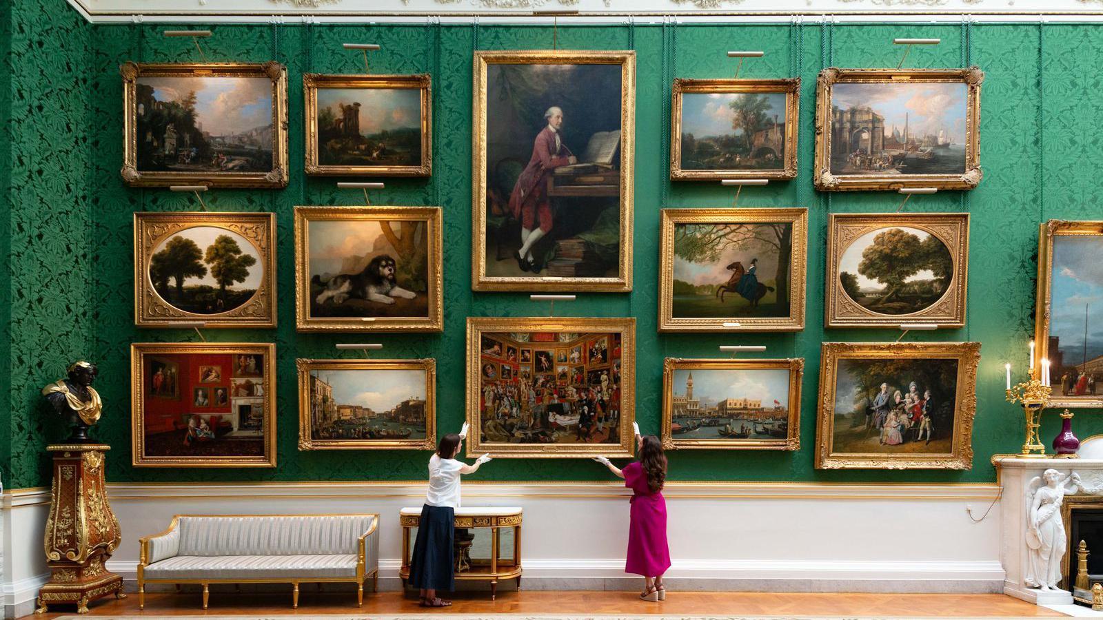 Dozens of paintings from the Royal Collection arranged on emerald-green silk walls. Two members of staff stand beneath the display, positioning The Tribuna of the Uffizi by Johan Zoffany among the newly rehung works.