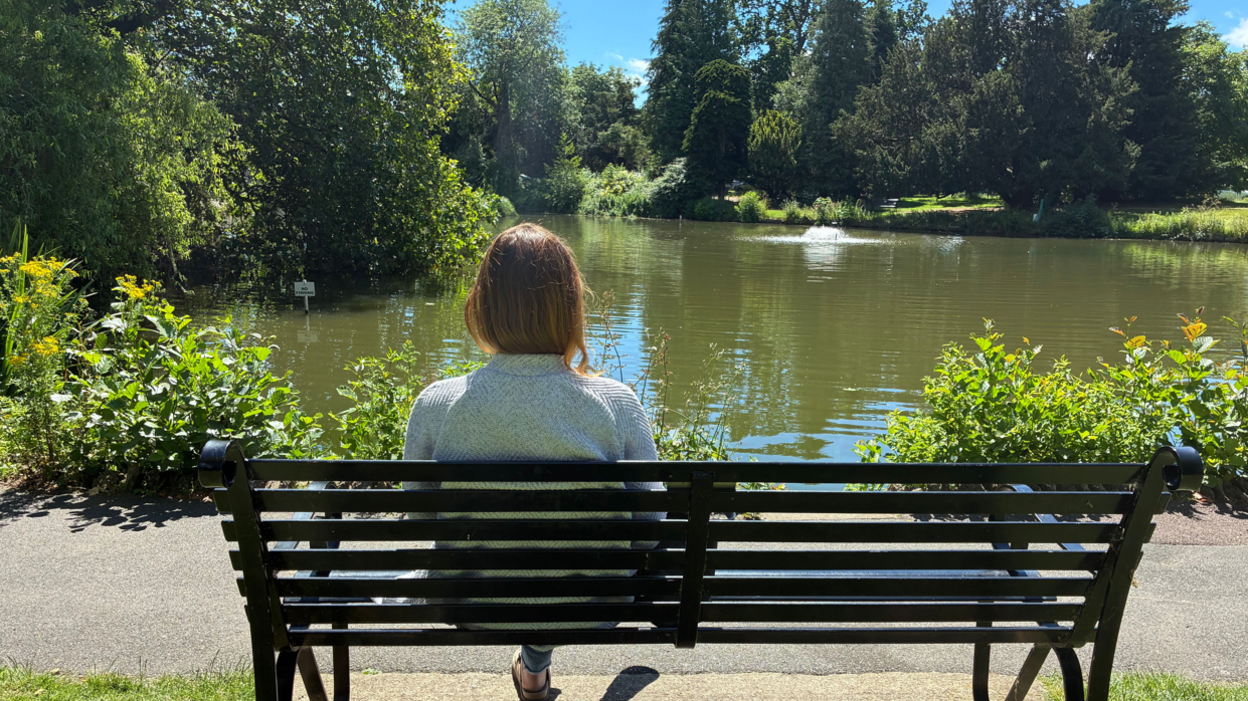 A woman is sat on a bench next to a lake. You can see the back of her body as she looks at the water. There are green plants around the water's edge, trees in the background and a fountain in the middle of the lake.