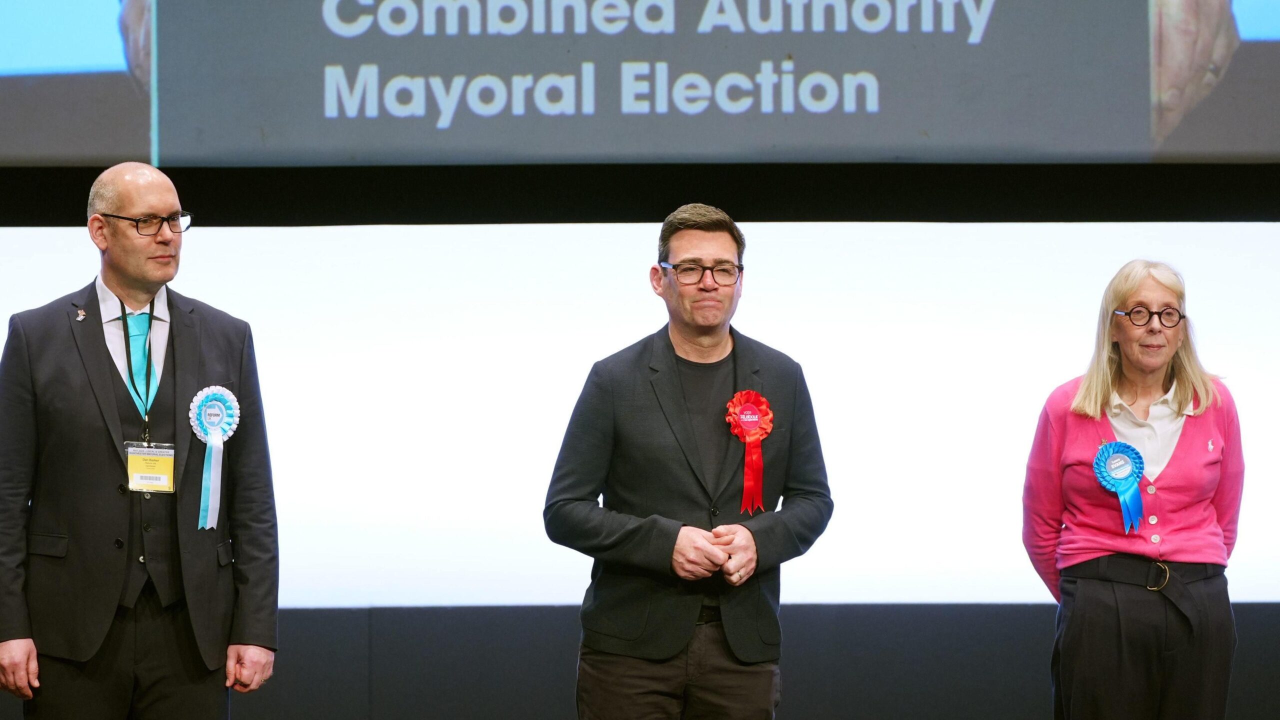 Andy Burnham (centre) is re-elected as Greater Manchester Mayor in 2024 as Reform candidate Dan Barker and Conservative candidate Laura Evans look on. Burnham wears a black jacket and trousers with a black t-shirt.
