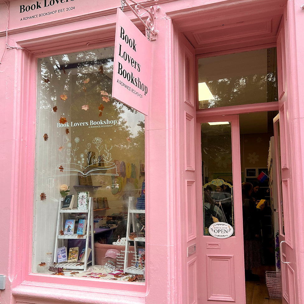 Book Lovers Bookshop from the outside. It has doors to the right and a window to the left and is painted in light pink. A sign also in pink with Book Lovers Bookshop A Romance Bookshop written in black hangs from a pink metal bracket. Two A-shaped small bookshelves with books are on display in the window and there are leaves attached to the inside. People can be seen inside the door reading books.
