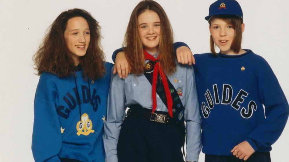 Three girls standing next to one another and smiling with their arms around each other. The girl on the left is wearing a blue hoodie with the word 'Guides' on it in navy text above a yellow and white Girlguiding logo. The girl in the middle is wearing a light blue shirt, red neckerchief, and badge sash. The girl on the right is wearing a royal blue sweatshirt with 'Guides' on it in large letters and a cap.