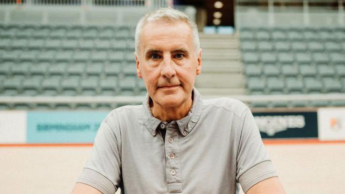 Dermot Murnaghan looks into the camera while wearing a grey t-shirt in an arena in Birmingham in 2022.