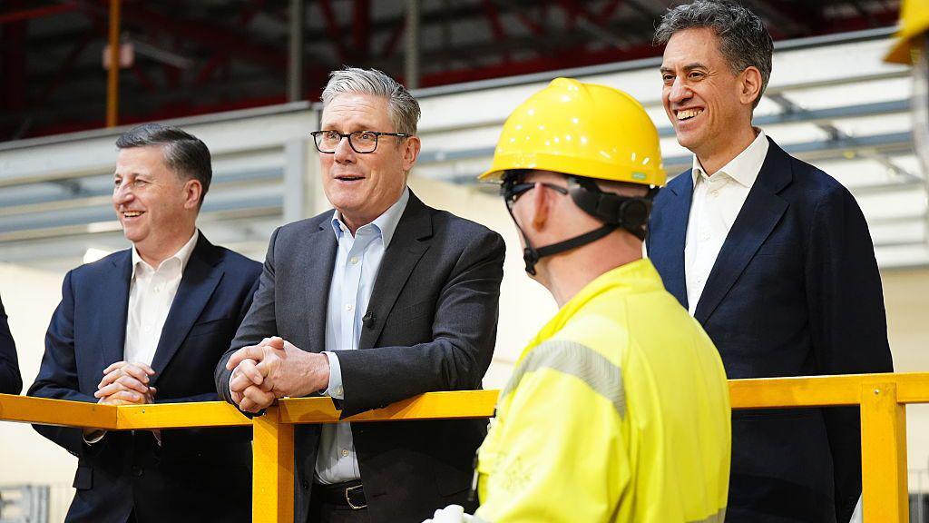 Two people in business attire stand on an elevated platform inside an industrial facility, speaking with a worker wearing a high-visibility jacket and yellow safety helmet in the foreground. Yellow safety railings and factory equipment are visible in the background.