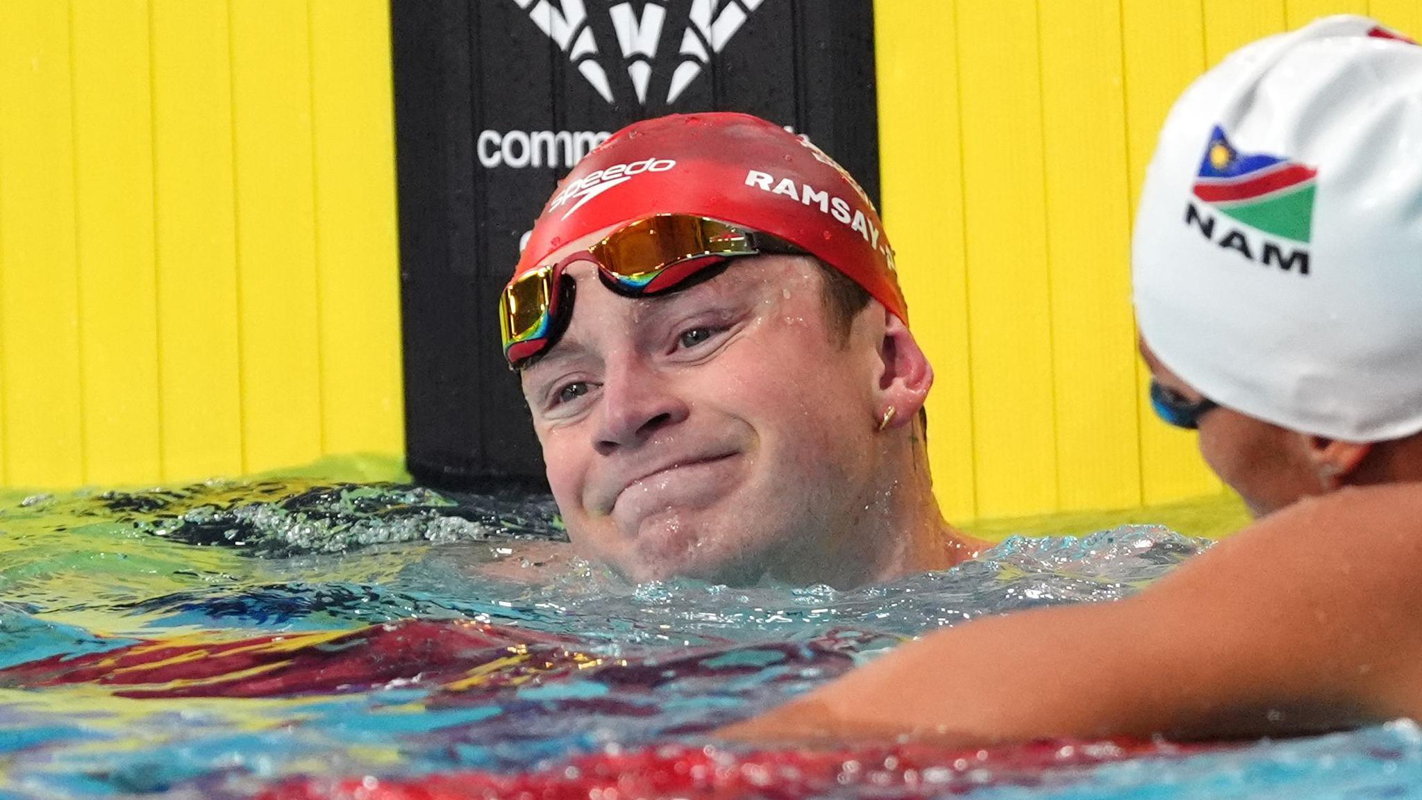 Adam Ramsay-Peaty with his head above water and goggles pulled up to his forehead as he wears a red swimming cap bearing his name