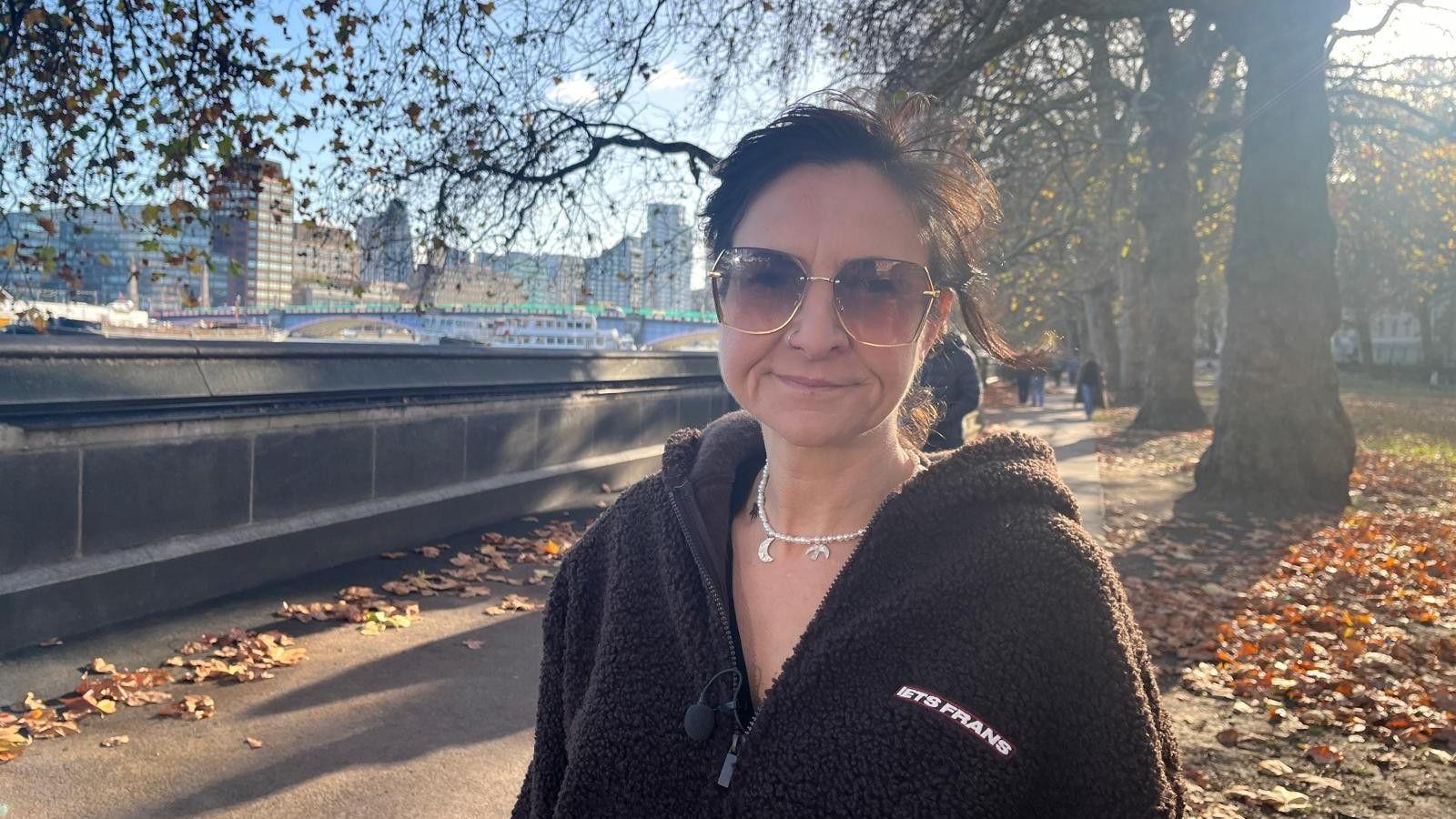 Jess Miller is standing next to the embankment in London. You can see the River Thames over her right shoulder and a row of trees directly behind her. She has short dark hair and wearing large brown sunglasses. She is wearing a silver necklace and a dark brown hoody