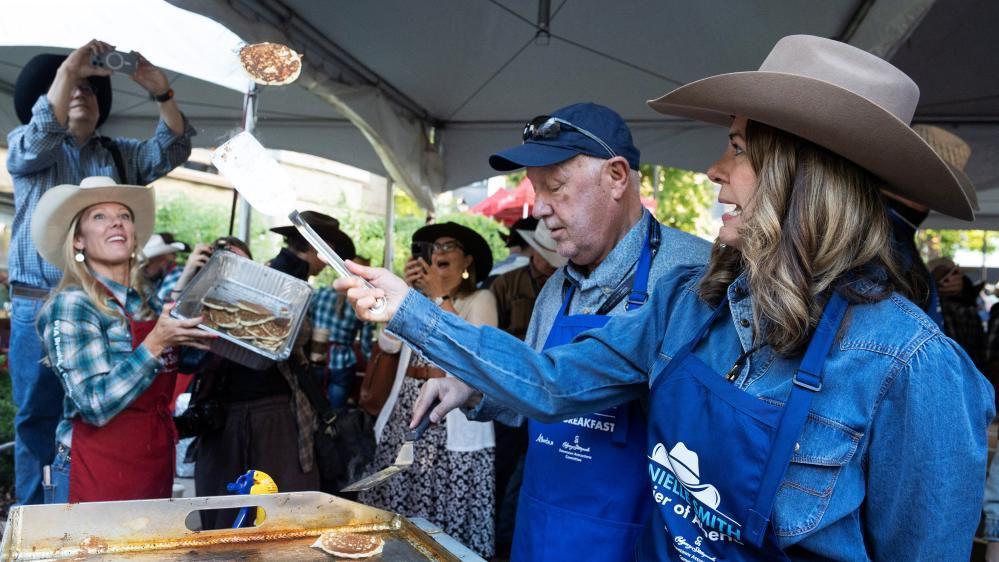 Alberta Premier Danielle Smith flips a pancake at the Premier's Stampede Breakfast. She is wearing a denim blue jean shirt and a cowboy hat, and has shoulder-length brown hair with blonde highlights.