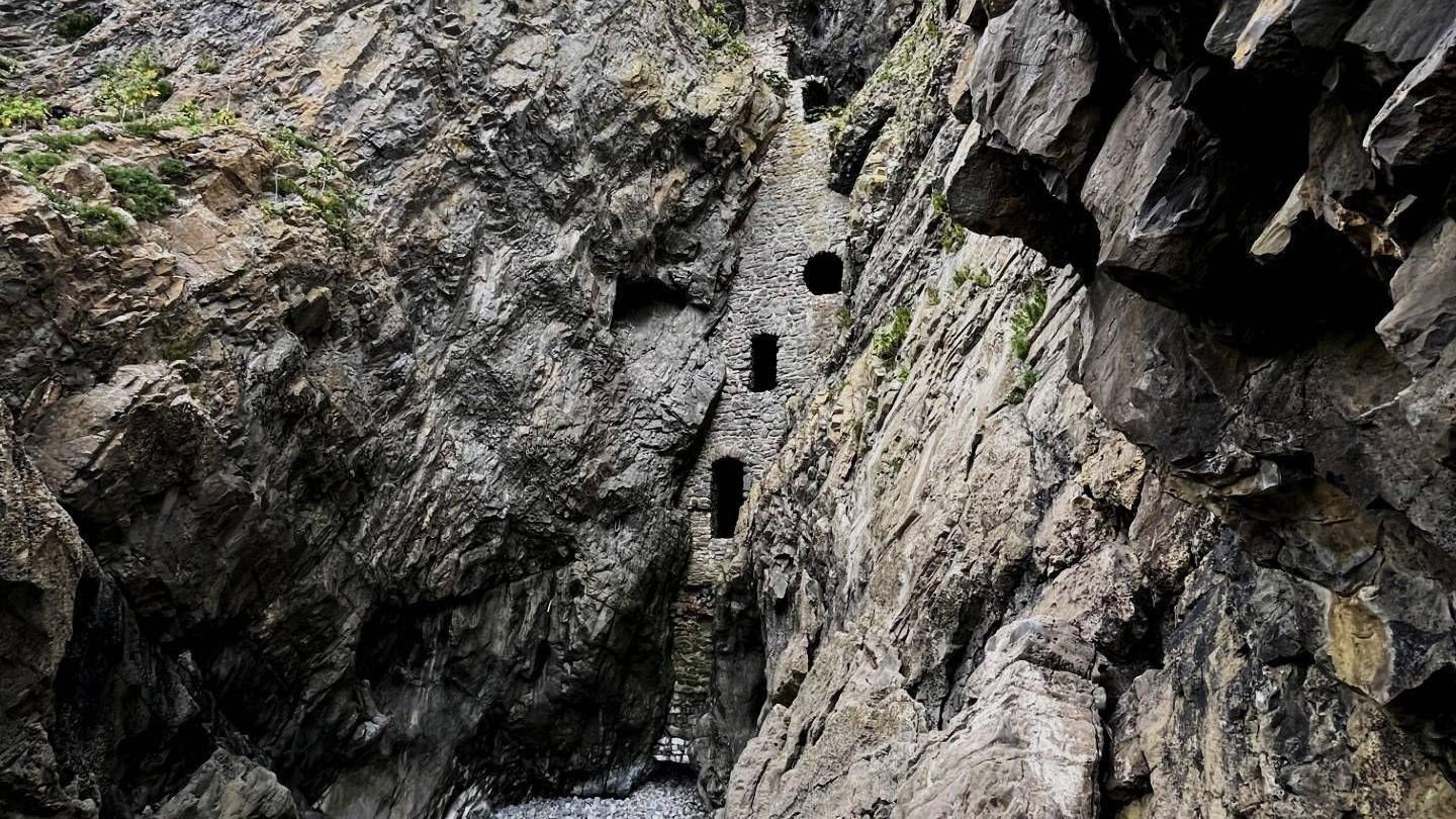 Wide view looking up through a rocky gorge with jagged grey cliff walls on both sides. A tall, narrow stone structure is embedded between the cliffs, with small window openings, and a dry, pebble-covered riverbed lies at the bottom.