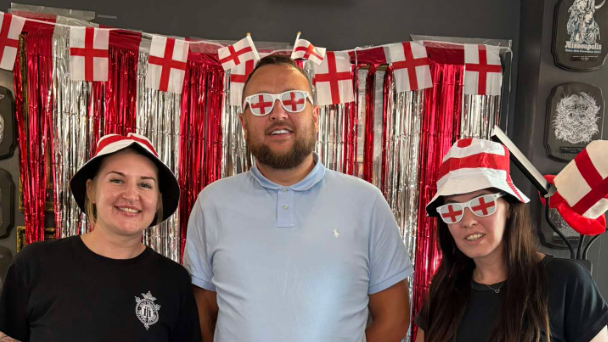 England football fans Amanda (left) Sean Carrington (centre) and Jamie Taylor (right) all dressed up with England red and white glasses and bucket hats. They are all smiling and standing in front of a shiny red and silver backdrop with England flags.
