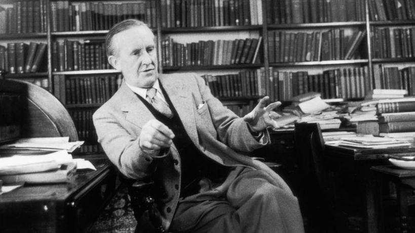 A black and white photograph of J.R.R. Tolkien sitting in a study, surrounded by bookcases and desks piled with books. He is wearing a suit and talking while making hand gestures.