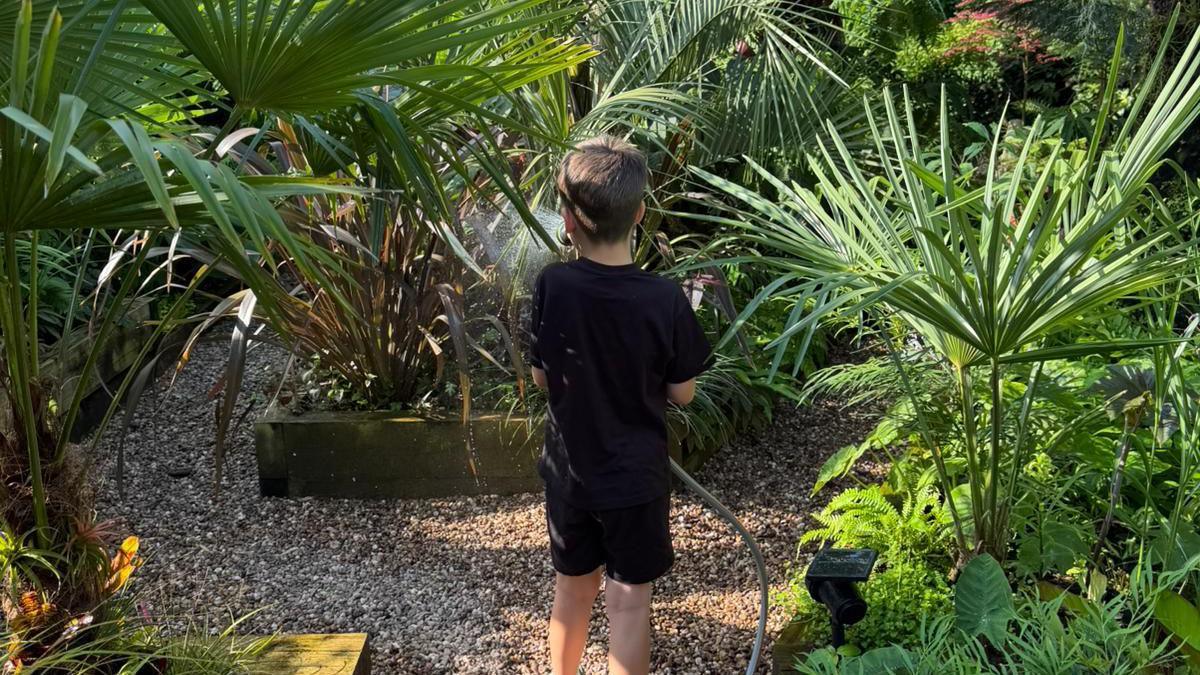 Dave and Gemma's son Arlo is photographed with his back facing the camera. He is wearing a black t-shirt with matching shorts. Arlo is holding a hosepipe and is watering the plants.