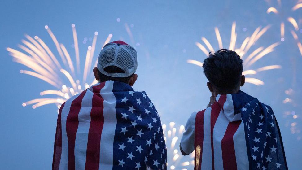 Two boys watch a fireworks display during a celebration of the US Army's 250th anniversary on Flag Day, June 14, 2025 in Washington DC. They are draped in USA flag and the sky in front of them is alight with fireworks