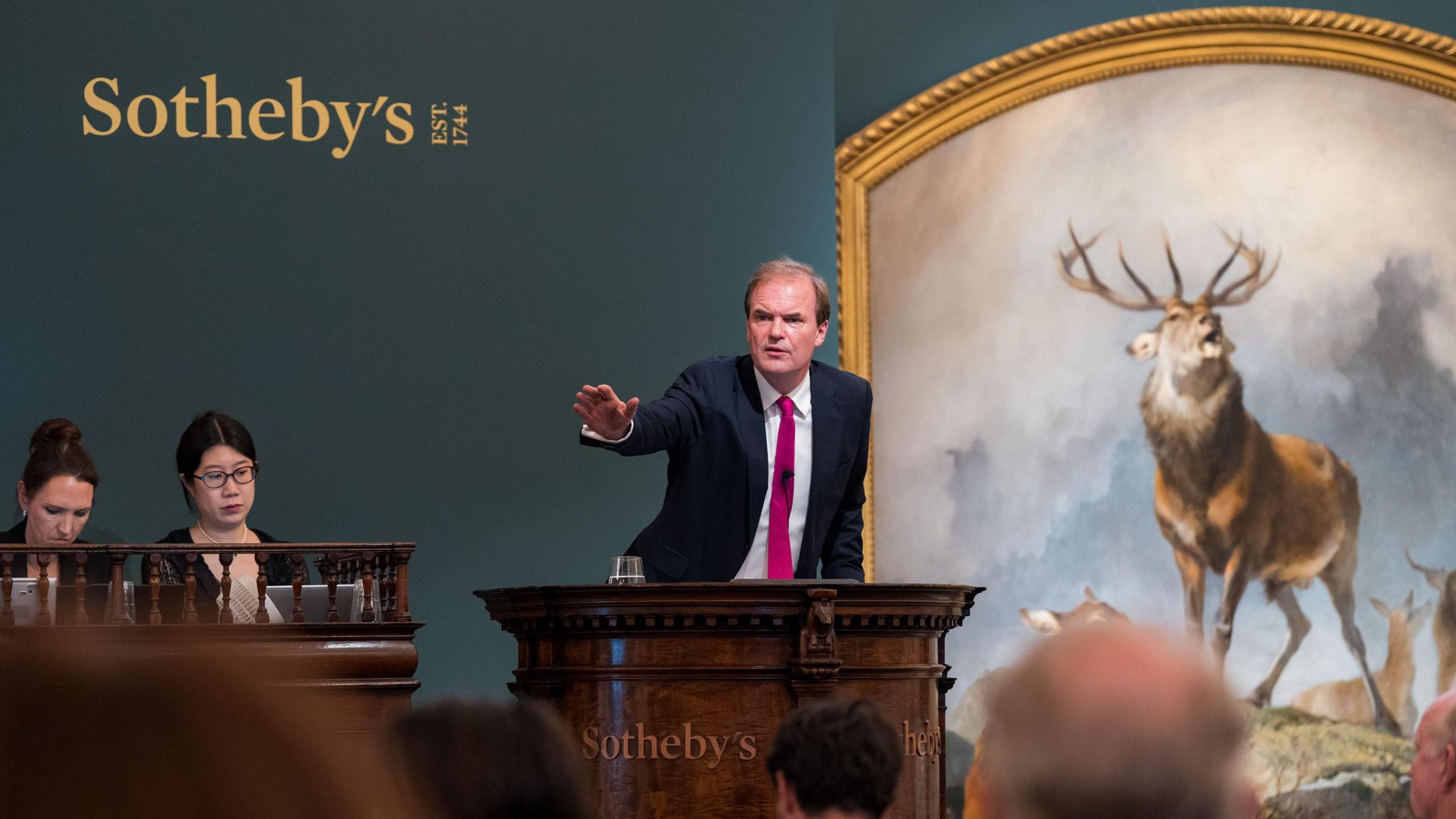 Auctioneer with his hand raised in front of the Scene of Braemar painting. Sothebys is written on the wall behind him