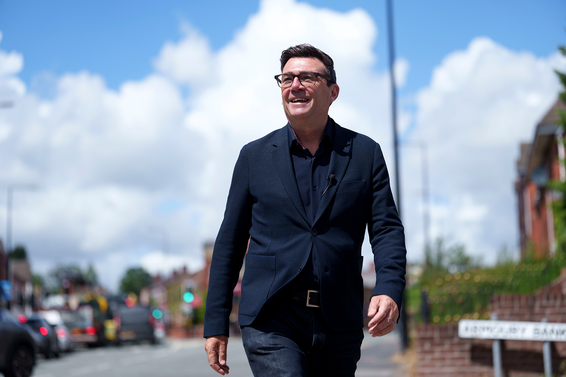 Andy Burnham smiles during a campaign visit to Ashton-in-Makerfield before the forthcoming by-election, in Manchester, England, Tuesday, June 9, 2026. He is wearing a dark suit jacket with a dark shirt underneath. He is smiling and waling in a sunny street. His hair is dark and he is wearing dark-framed glasses.