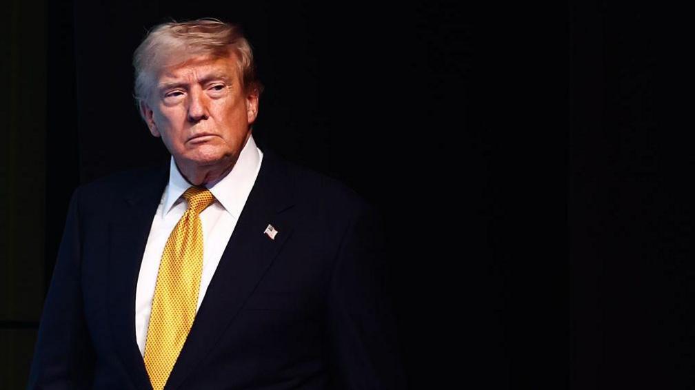 Trump seen wearing a suit with a yellow tie. The background is black