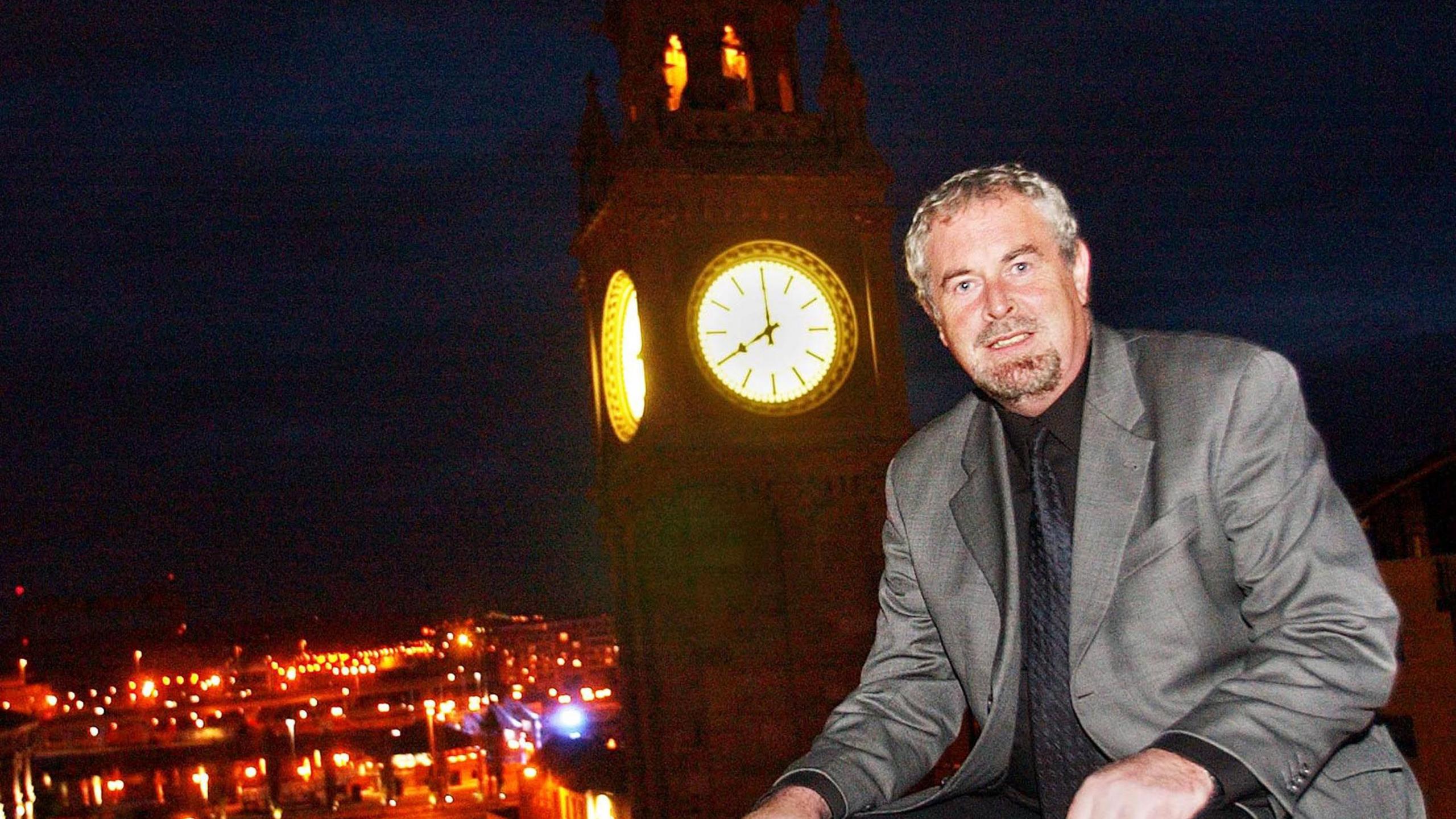 A man with short brown and grey hair wearing a grey suit as he sits in front of a lit up clock tower.
