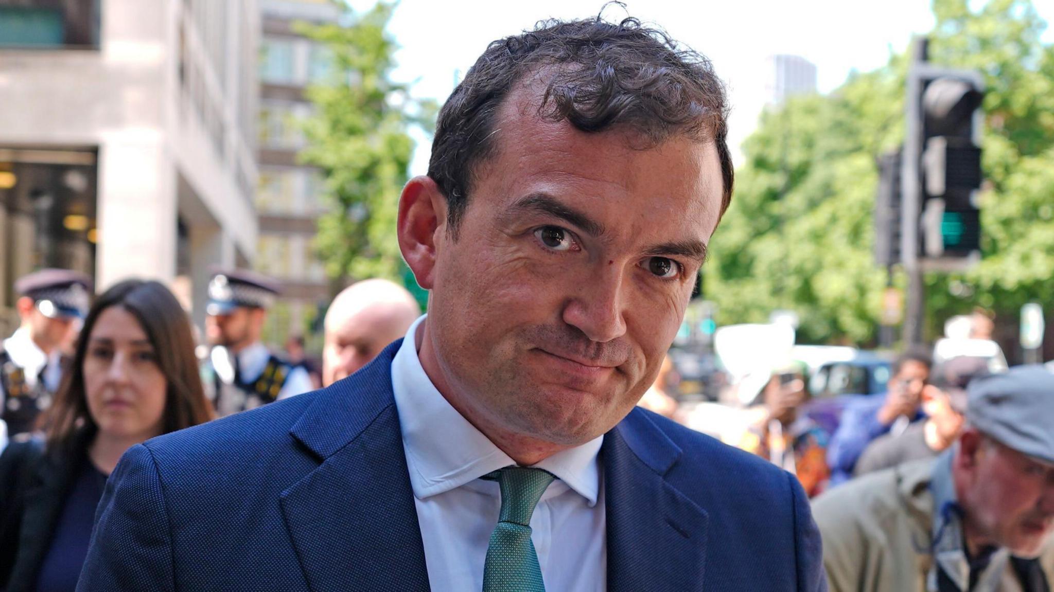 Patrick Spencer leaves a court, dressed in a blue suit and green tie. He is smiling at the cameras. In front of him is a police officer, dressed in uniform.