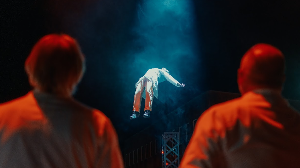 Two men with their backs to the camera watch as another man, in a white lab coat, who is floating in the air on his back with a light on him and smoke surrounding him.