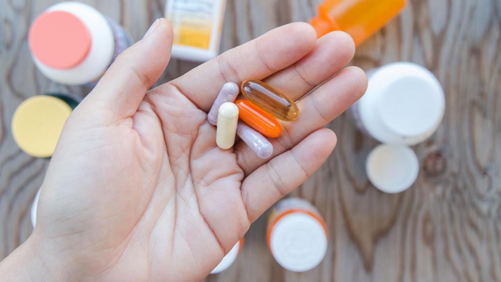 A hand holding a bunch of supplements in an open palm. There are bottles of supplements as a background.