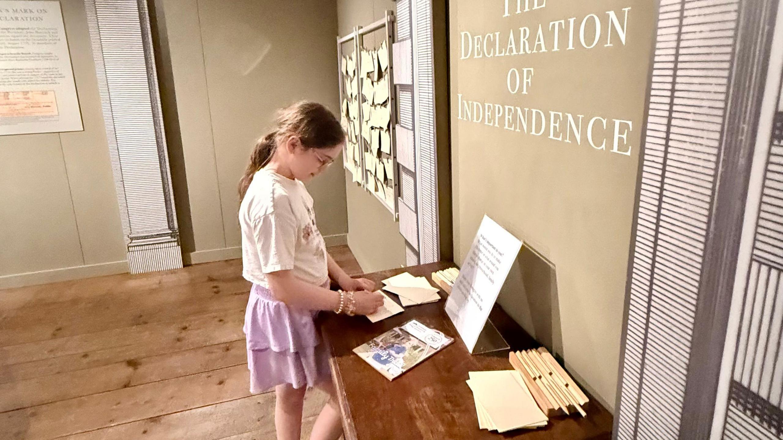 A girl writes on a piece of paper next to a display that reads "Declaration of Independence"