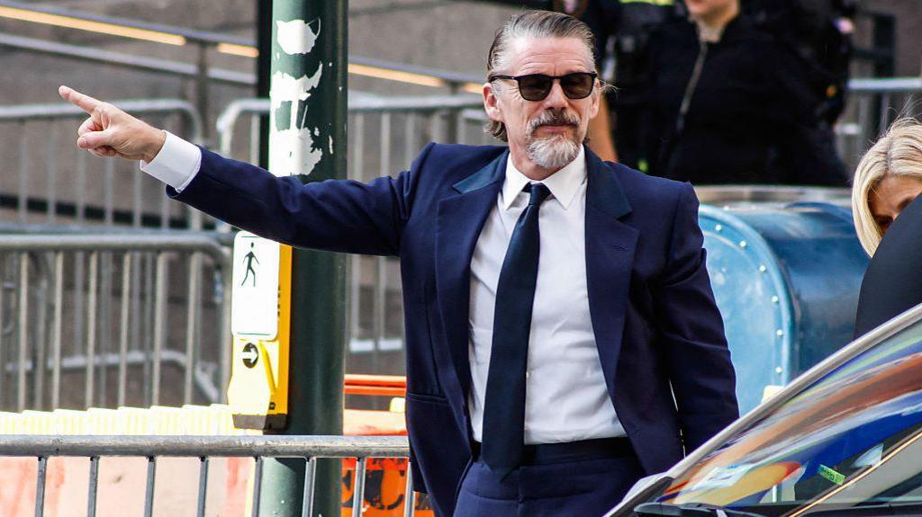 Ethan Hawke pictured wearing a navy suit. His arm is outstretched and his finger is pointed