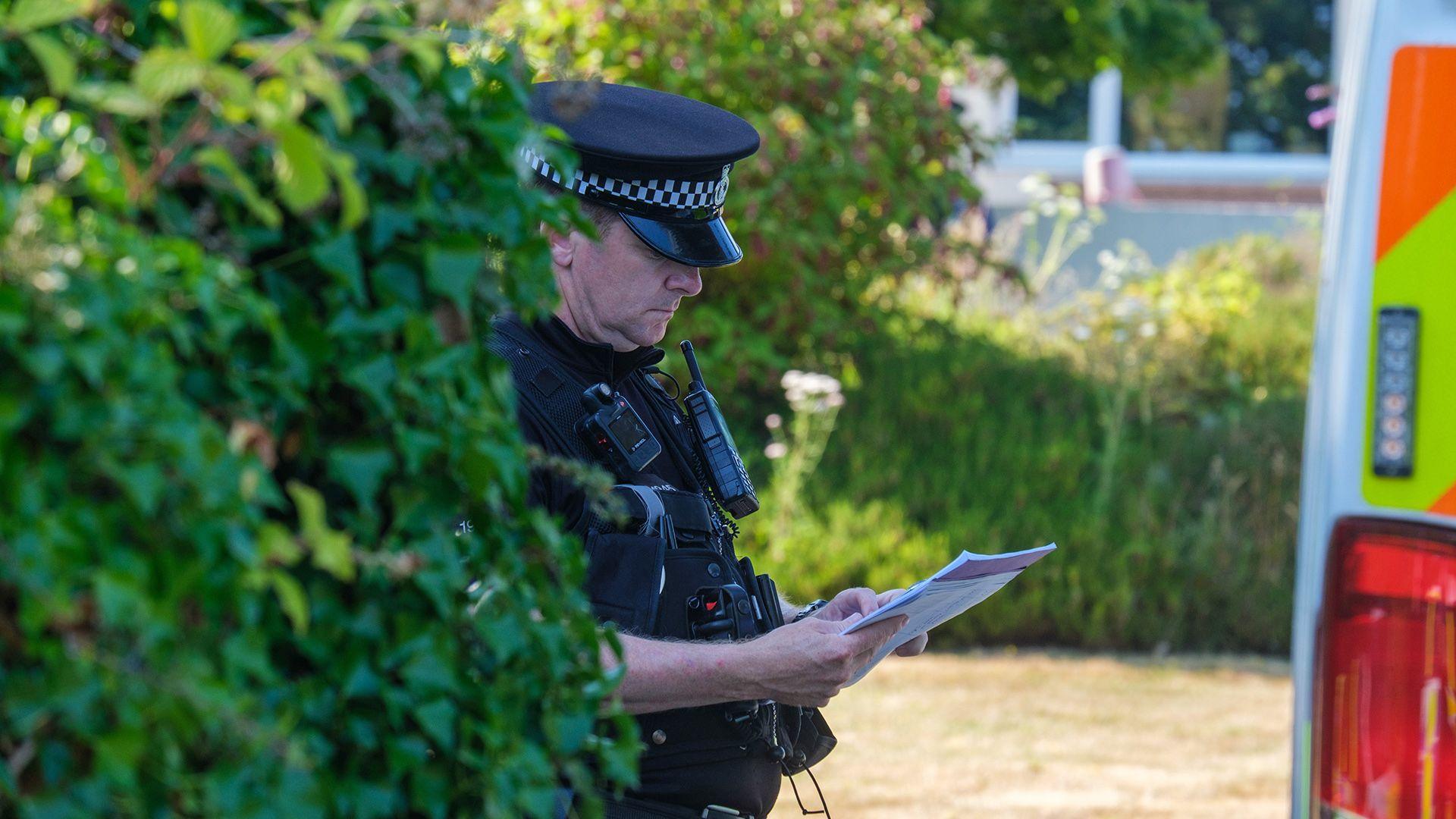 A police officer looks down at a sheert of paper while wearing uniform next to a bush and police van outside Widdecombe's home on Friday.
