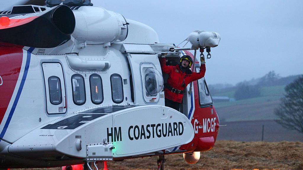A man in a red bodysuit and helmet leaning out the side of a HM Coastguard helicopter in white and red.
