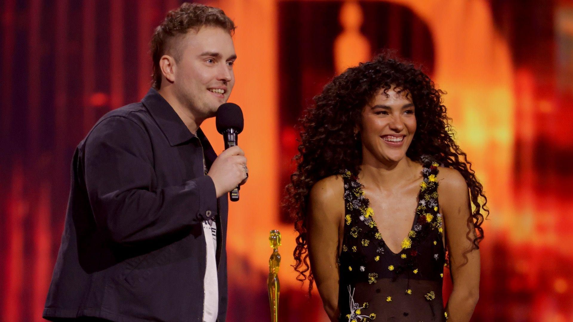 Sam Fender and Olivia Dean stand on a warmly lit stage. Sam is holding a microphone, whilst Olivia stands next to him, beside a golden statuette BRIT award.