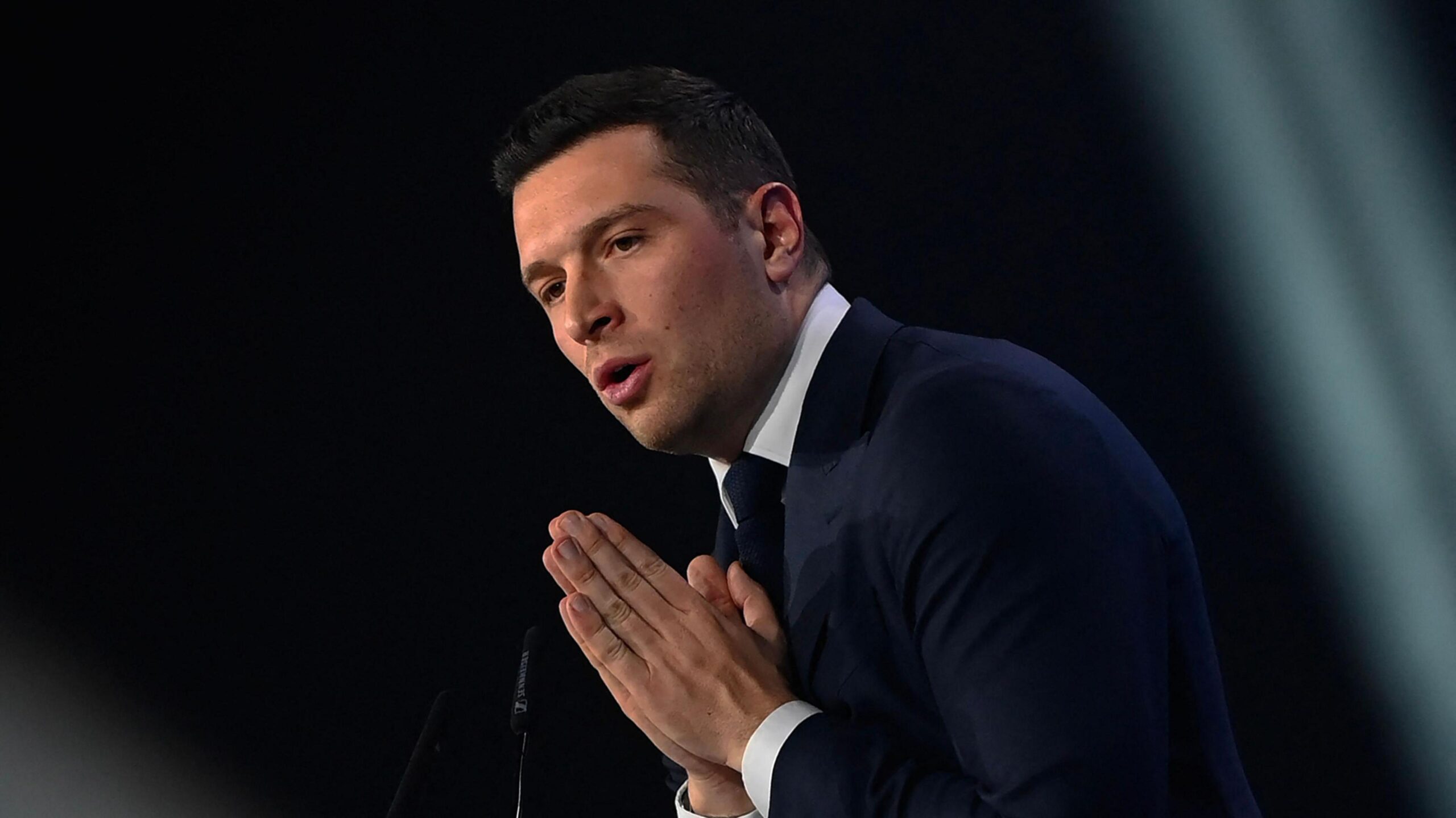 Jordan Bardella wears a suit and stands in front of a dark background with his palms pressed together.