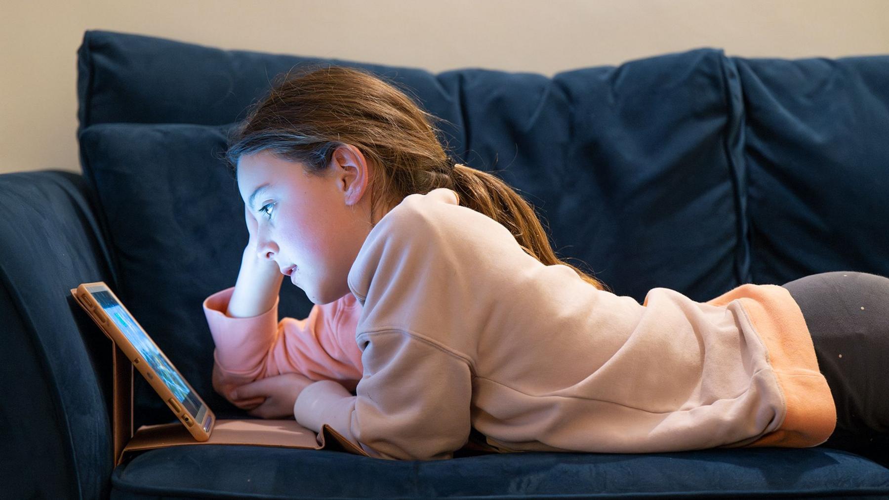 A young girl wearing a cream top and grey leggings is lying on a sofa staring at a brightly lit screen in a dark room