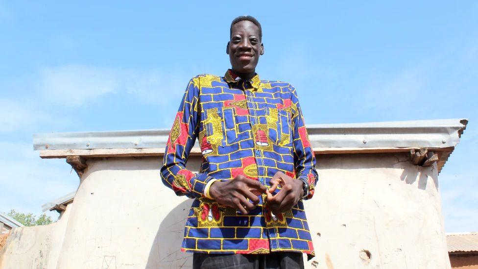 A smiling Sulemana Abdul Samed, also known as Awuche, stands in front a a building, towering over its roof. He wears a colourful shirt.