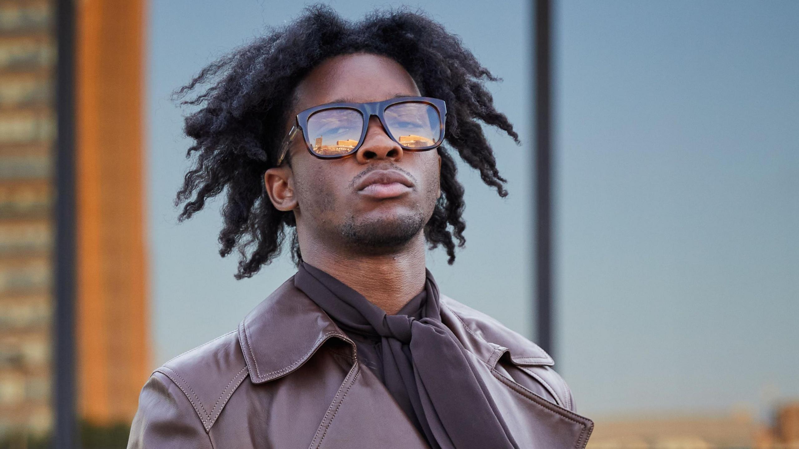 D4vd is pictured in large, black-rimmed glasses and a brown leather coat