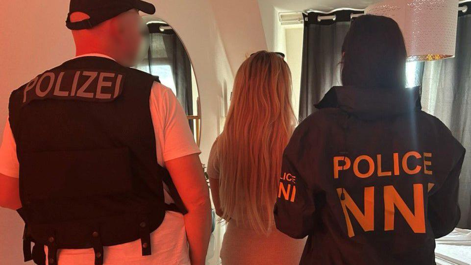 A woman is pictured standing in between two police officers with her back turned. The officers are also facing away. A male officer's face has been blurred.