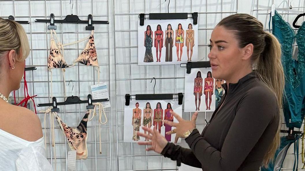 Olivia wearing a brown long sleeve top talking to another woman in front of a metal grid covered in bikinis and design sketches.