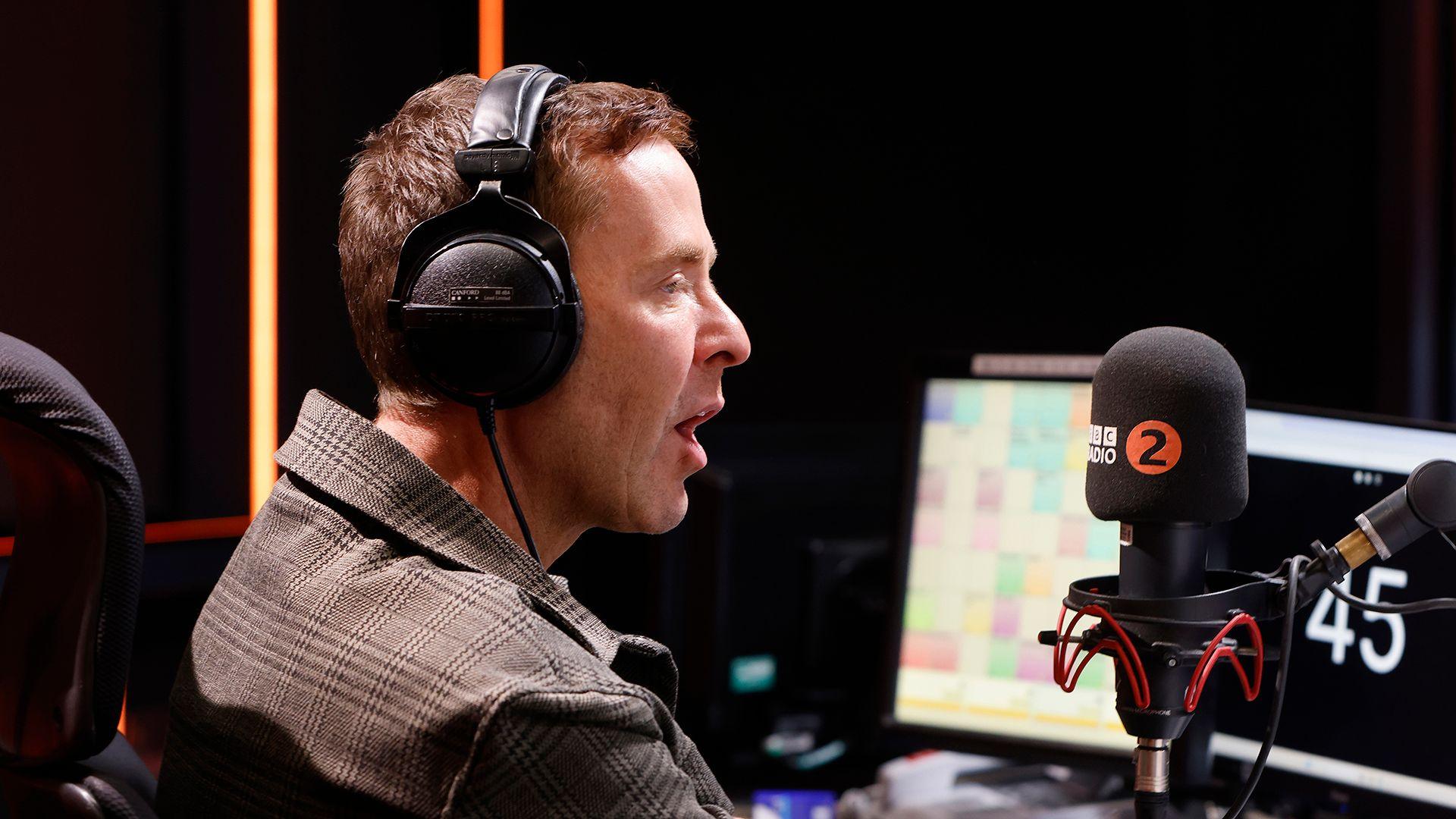 A profile image of Scott Mills wearing headphones while speaking into a microphone with the BBC logo and the number 2 on it, with computer screens and the studio in the background, in early 2026.