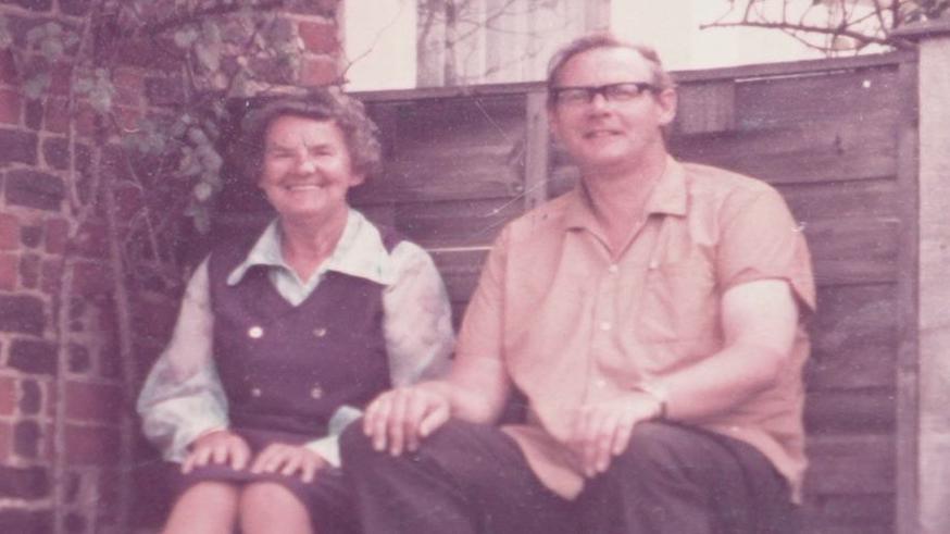 A faded colour photograph of Anthony Littler and an older woman sitting side by side on a bench in front of a brick wall and wooden fence. He wears glasses and a short-sleeved shirt; she wears a dark pinafore over a white blouse. Both are smiling.