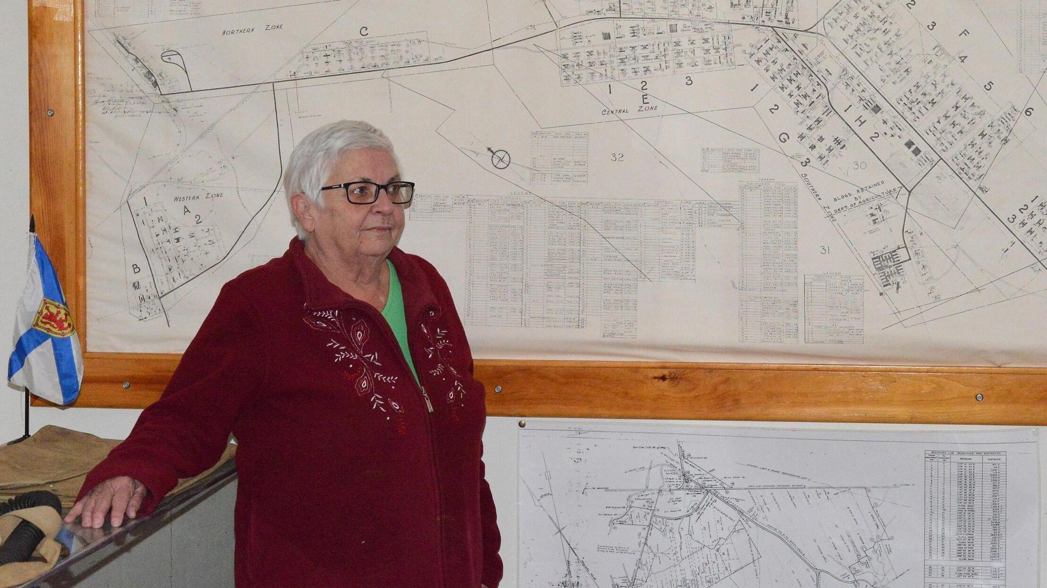 Annette Sharpe, stands to the left and looks away from the camera. She wears a maroon cardigan over a bright green shirt, and has dark-rimmed square glasses and short white hair. Behind her on the wall are black and white maps