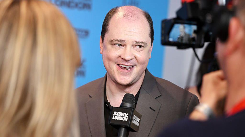 Mike Flanagan pictured mid-interview on a red-carpet. He's wearing a dark brown suit jacket over a black t-shirt. The camera focuses on him over the shoulders of a reporter and cameraman.