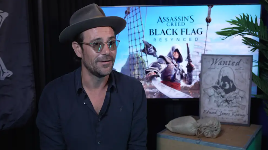 Matt Ryan is sitting on a chair wearing a hat and glasses, speaking to someone on the other side of the camera. Behind him is promotional material for the upcoming Assassin's Creed Black Flag game.