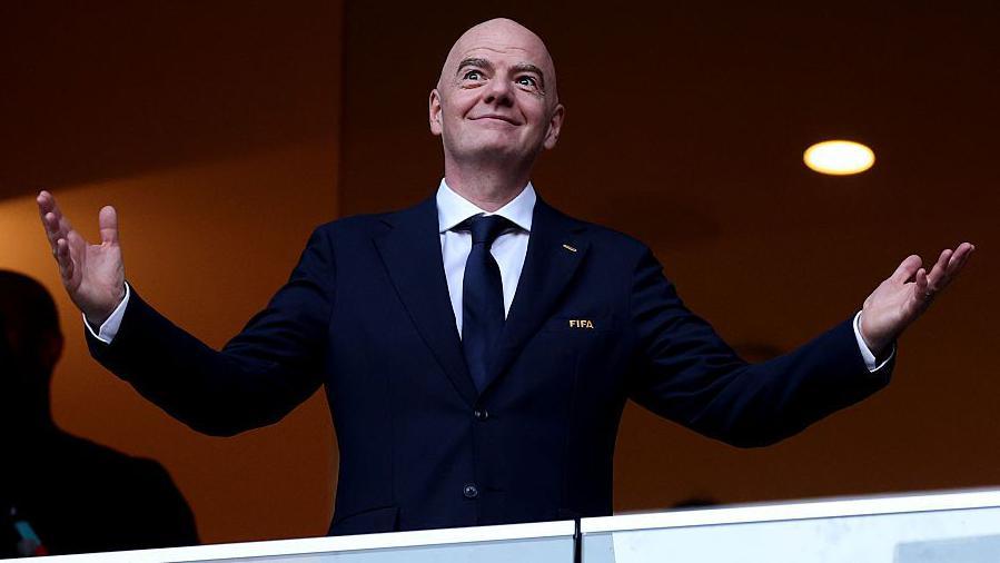 Gianni Infantino holds his arms out at each side with open palms, as he smiles while wearing a buttoned-up, dark smart jacket with FIFA written on its breast pocket