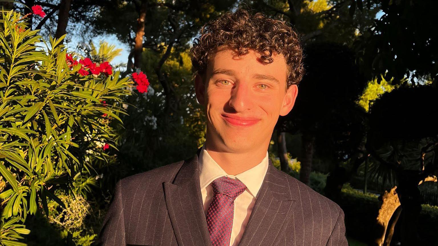 Daniel, who has dark curly hair and is wearing a purple suit stood outside with green bushes and red flowers in the background.