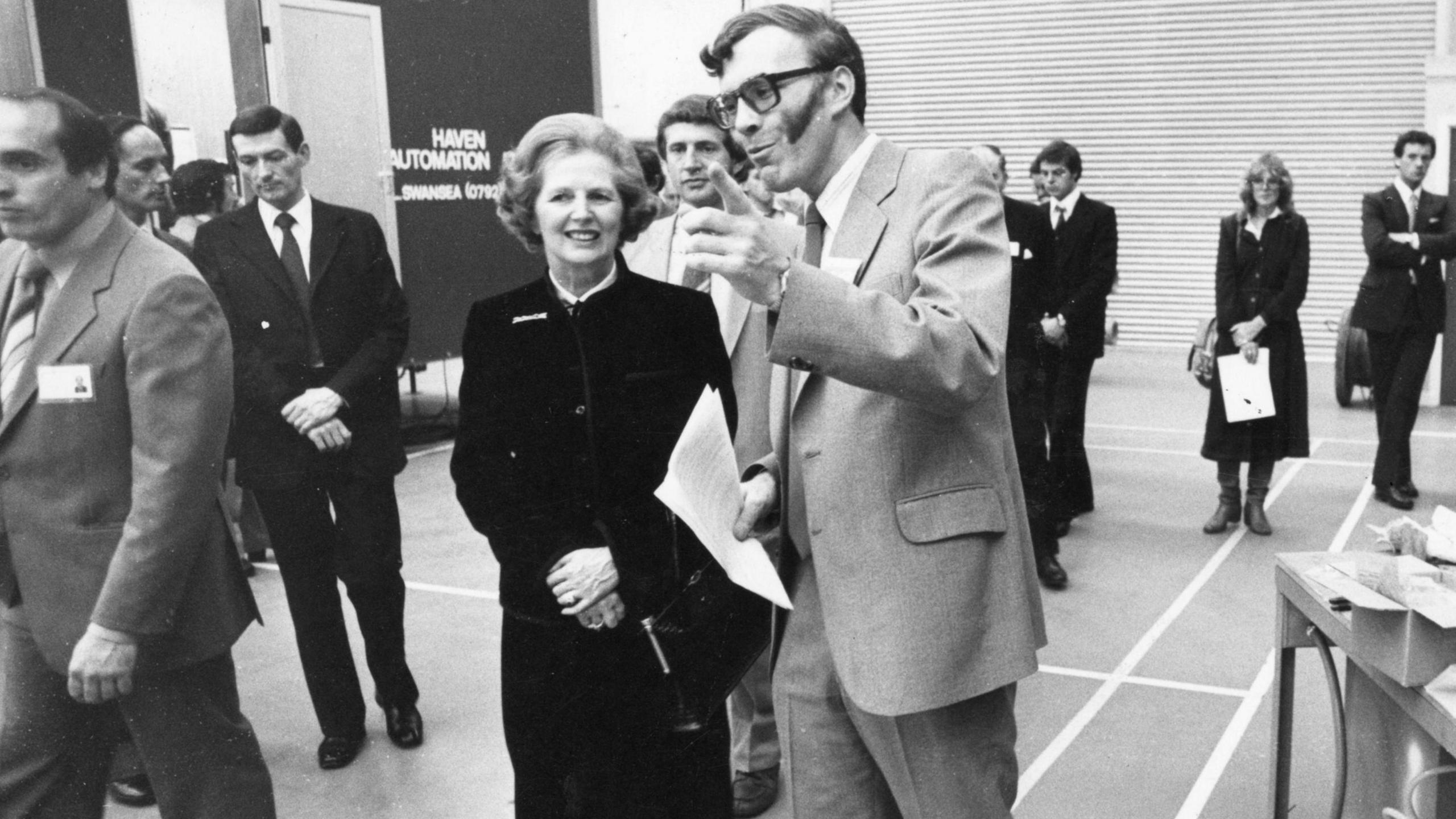 A black and white image of Sir Ian Wood, in light suit, showing Margaret Thatcher around a hall. He is pointing and she is smiling