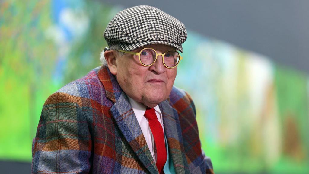 David Hockney, wearing round glasses and a flat cap, along with a multicoloured checked shirt, in front of an artwork that can be seen blurred in the background
