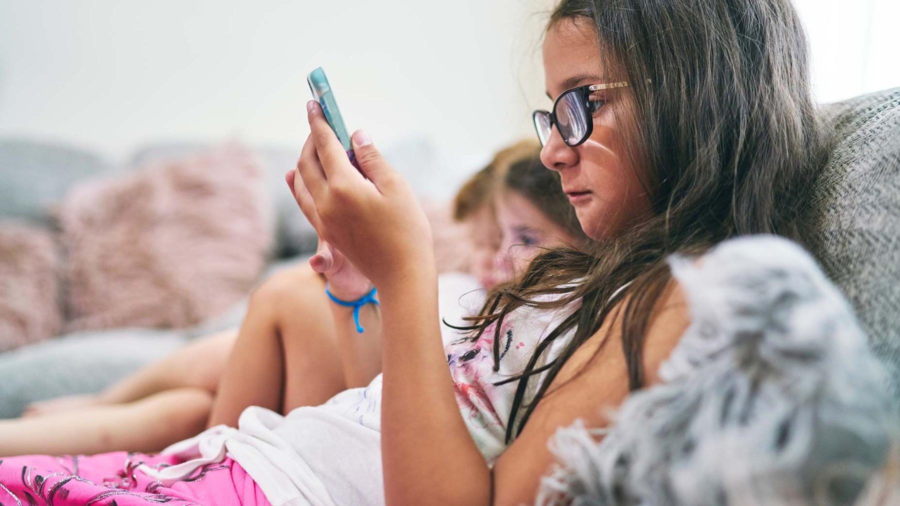 A young girl wearing pink and white is sitting on a sofa looking at her phone - in the background are two other childrenalso looking at a screen