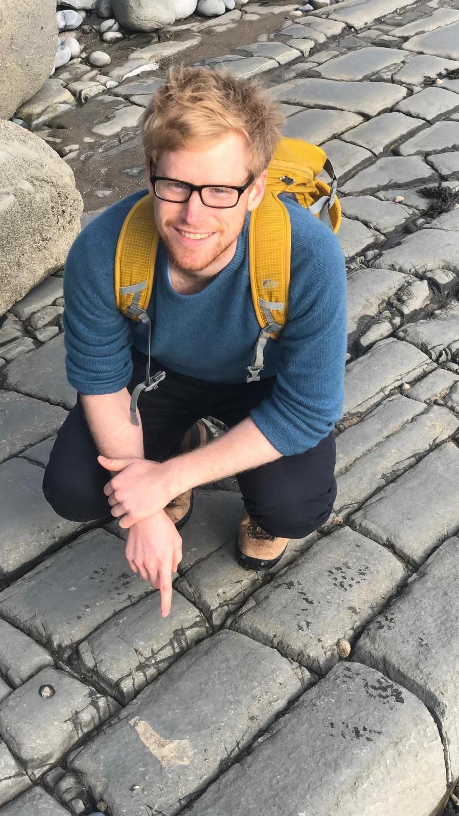 Matthew, wearing dark rimmed glasses and a yellow backpack, standing on rocky ground pointing to a fossil on the stones in front of him.