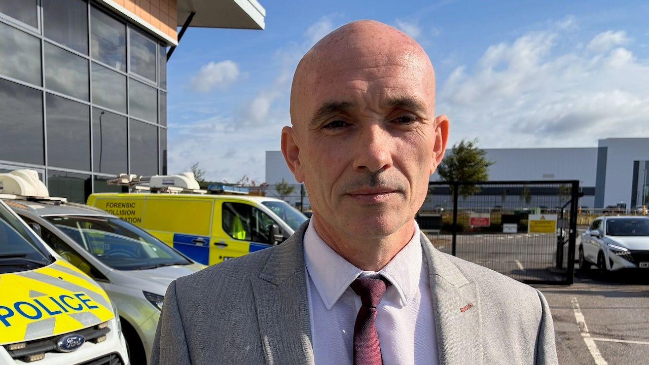 Detective Superintendent Al Curtis is dressed in a grey suit with a burgundy tie. He is stood in front of three police vehicles in a car park. He is looking at the camera with a sombre expression.