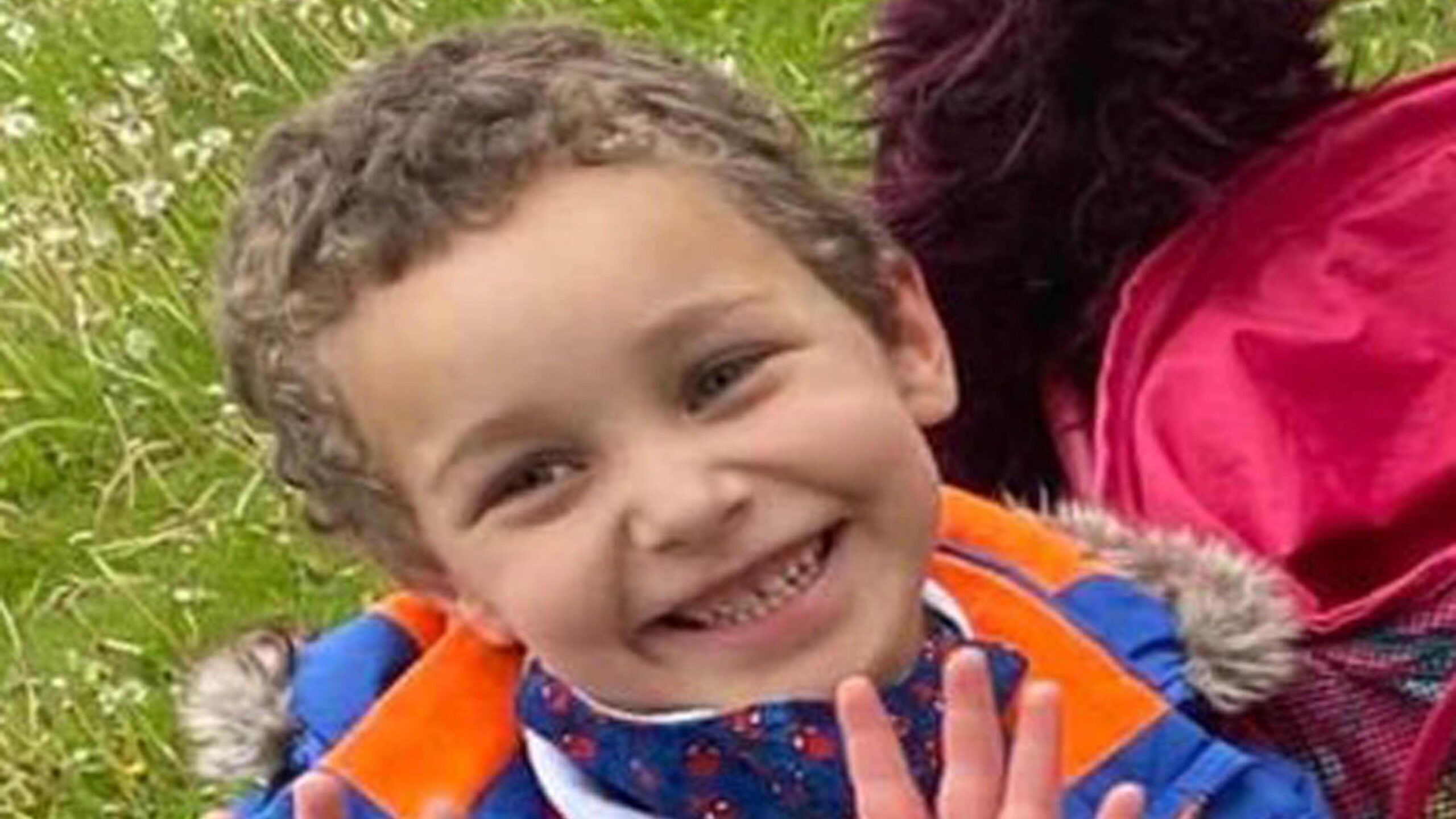 A little boy with curly brown hair. He's outside surrounded by grass and wearing a blue and orange coat.