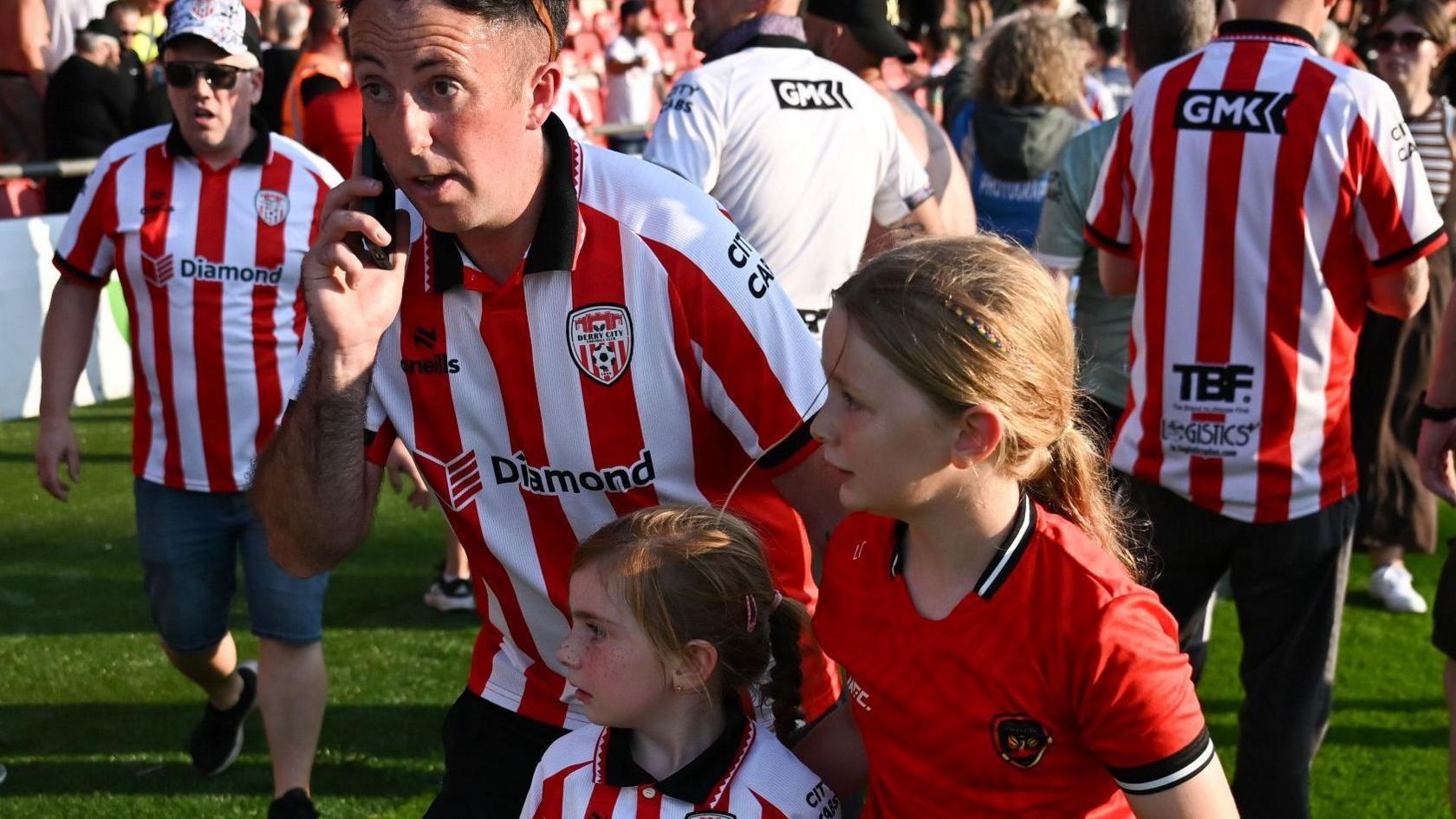 Two girls in Derry City tops look fearfully out of frame as a young man with them talks on the phone. There are crows of people around them, standing on a football pitch